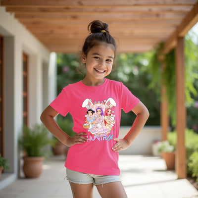 Young girl wearing a pink t-shirt with colorful kpop huntrix graphics and text, standing outdoors.