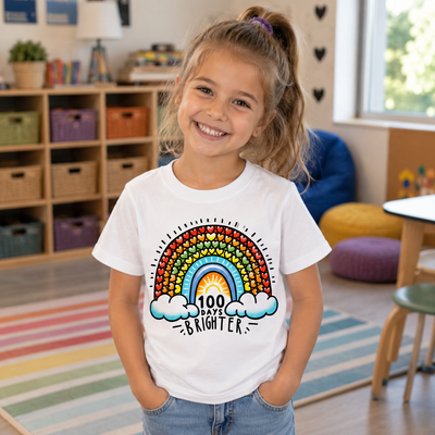 A young girl smiling in a colorful children's room, wearing a white graphic t-shirt with a rainbow design and the text '100 DAYS BRIGHTER'.