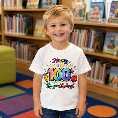 A young boy smiling in a children's bookstore, wearing a white t-shirt with a colorful 'Happy 100th Day of School' graphic print.