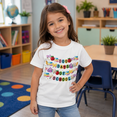 A young girl with a pink hair bow stands in a classroom wearing a white graphic t-shirt that reads '100 DAYS OF SCHOOL' in colorful letters with school-themed illustrations. She is smiling and posing with one hand on her hip.