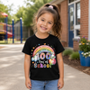 A young girl smiling outdoors in front of a brick school building, wearing a black short-sleeve T-shirt with a colorful '100th Day of School' rainbow graphic on the front.