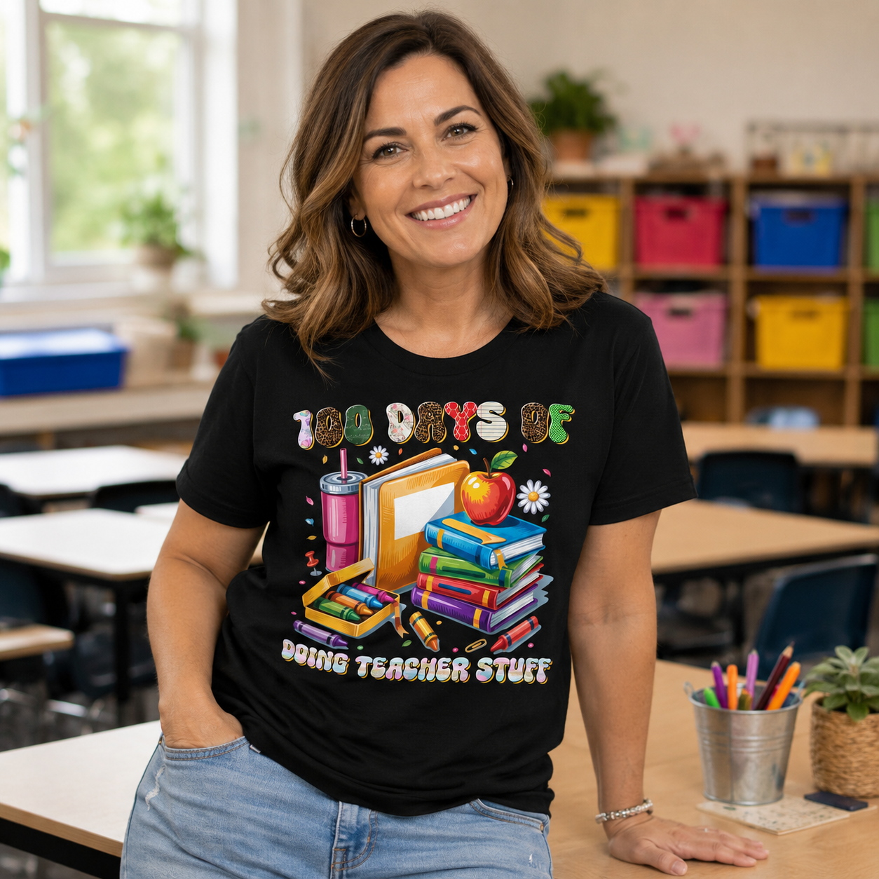 A smiling woman wearing a black graphic t-shirt that reads '100 DAYS BE DOING TEACHER STUFF' with colorful school supplies and an apple illustration, standing in a bright classroom setting.