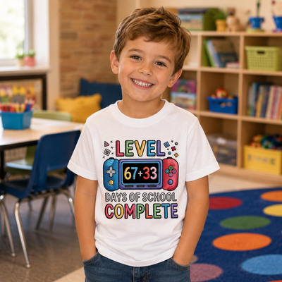 A young boy smiling in a colorful classroom, wearing a white graphic t-shirt that reads 'LEVEL DAYS OF SCHOOL COMPLETE' with a retro game controller design.
