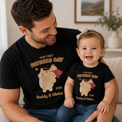 A smiling man and toddler daughter wearing matching black graphic T-shirts in a bright living room. The shirts feature a cartoon bear wearing a crown and cape with the text 'OUR FIRST FATHERS DAY 2026 Daddy & Me' in tan lettering.