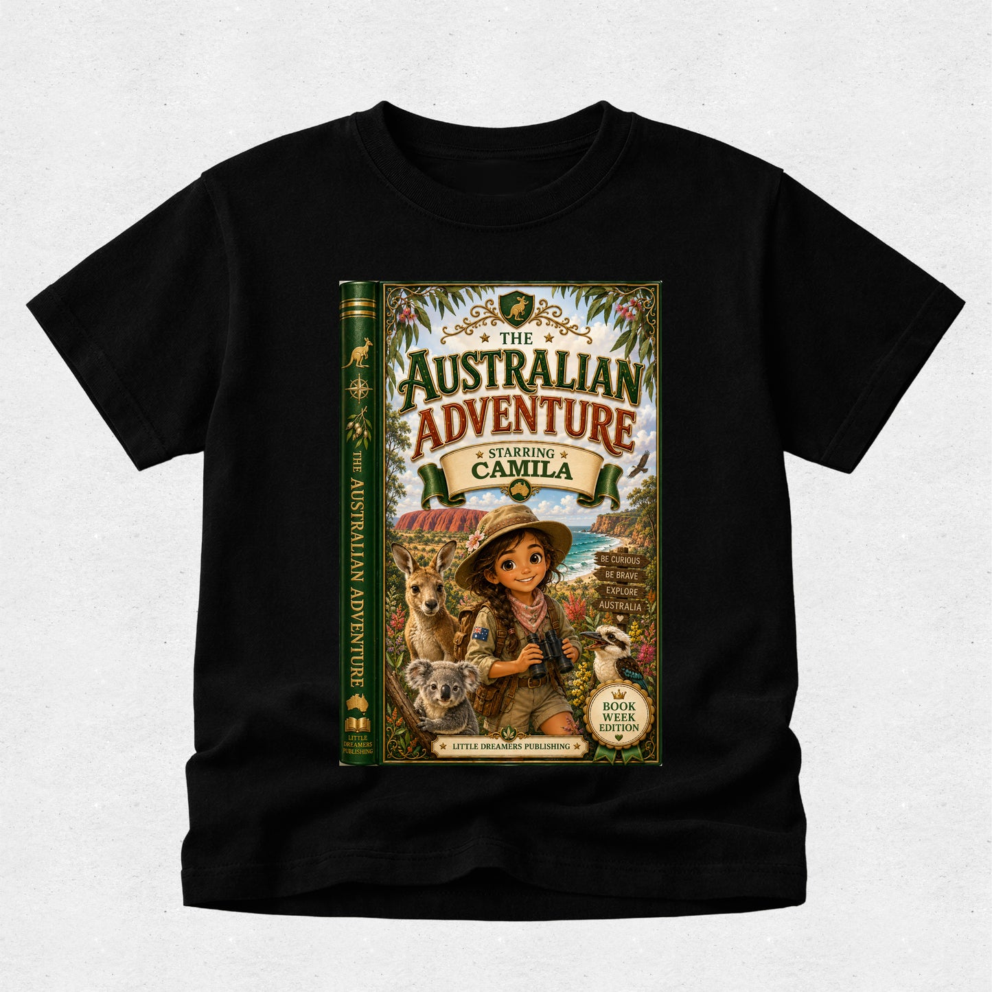 A black short-sleeve t-shirt with a large graphic print on the front depicting a book cover titled 'The Australian Adventure: Starring Camila'. The graphic features an illustration of a young girl in a safari outfit with a koala and a kangaroo, set against a light gray textured background.