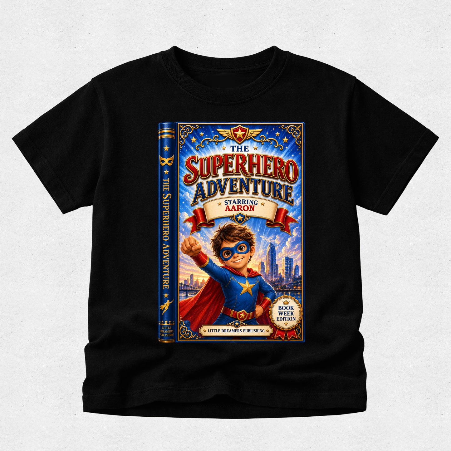 A black short-sleeve crewneck t-shirt featuring a large graphic print of a children's book cover titled 'The Superhero Adventure Starring Aaron'. The graphic shows a cartoon boy superhero in a blue and red costume with a city skyline in the background.