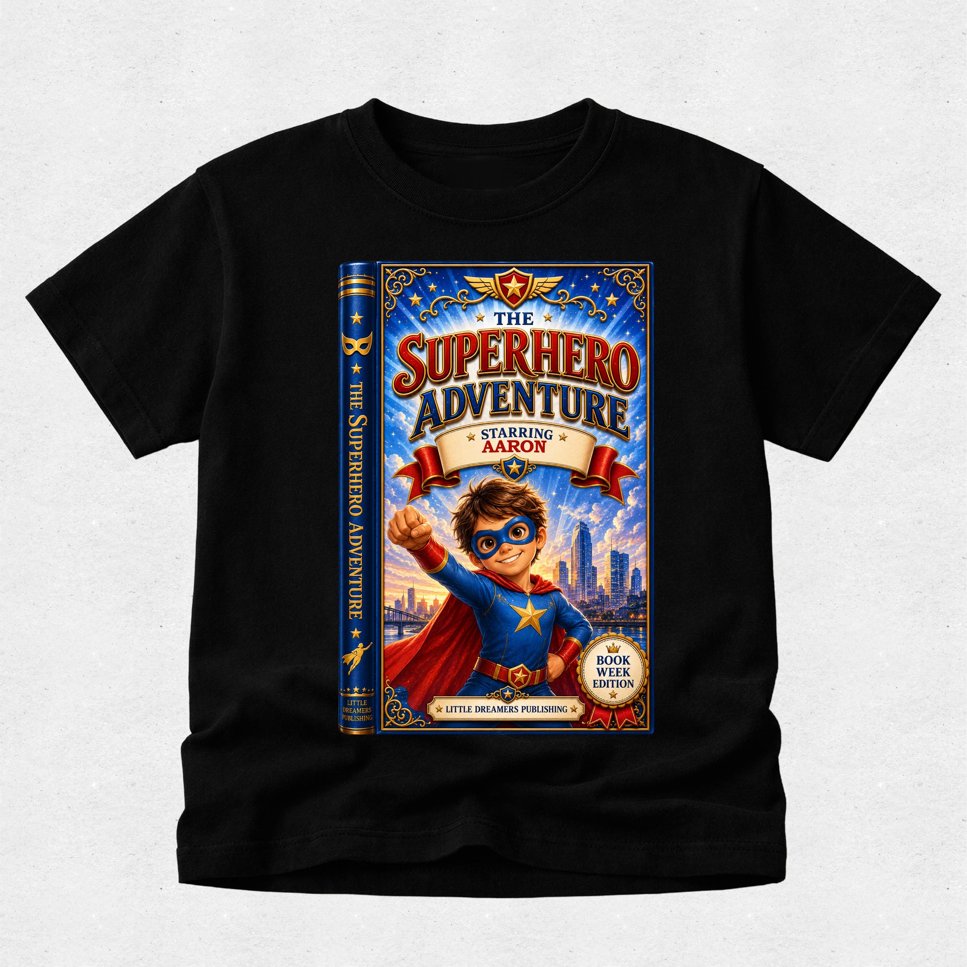 A black short-sleeve crewneck t-shirt featuring a large graphic print of a children's book cover titled 'The Superhero Adventure Starring Aaron'. The graphic shows a cartoon boy superhero in a blue and red costume with a city skyline in the background.