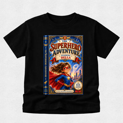 A black short-sleeve crewneck t-shirt featuring a large graphic print of a book cover titled 'The Superhero Adventure' with an illustration of a girl in a superhero costume, displayed on a light gray textured background.