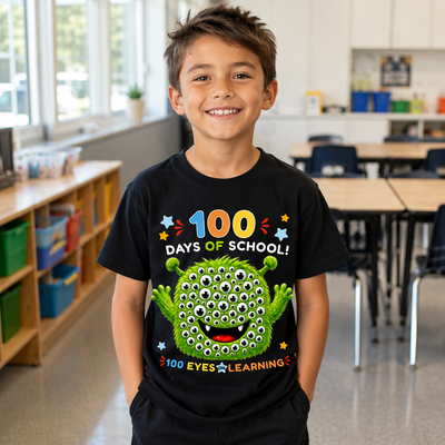 A young boy smiling in a bright classroom, wearing a black graphic t-shirt that reads '100 DAYS OF SCHOOL!' and features a green monster with many eyes. The text '100 EYES LEARNING' is printed below the graphic.