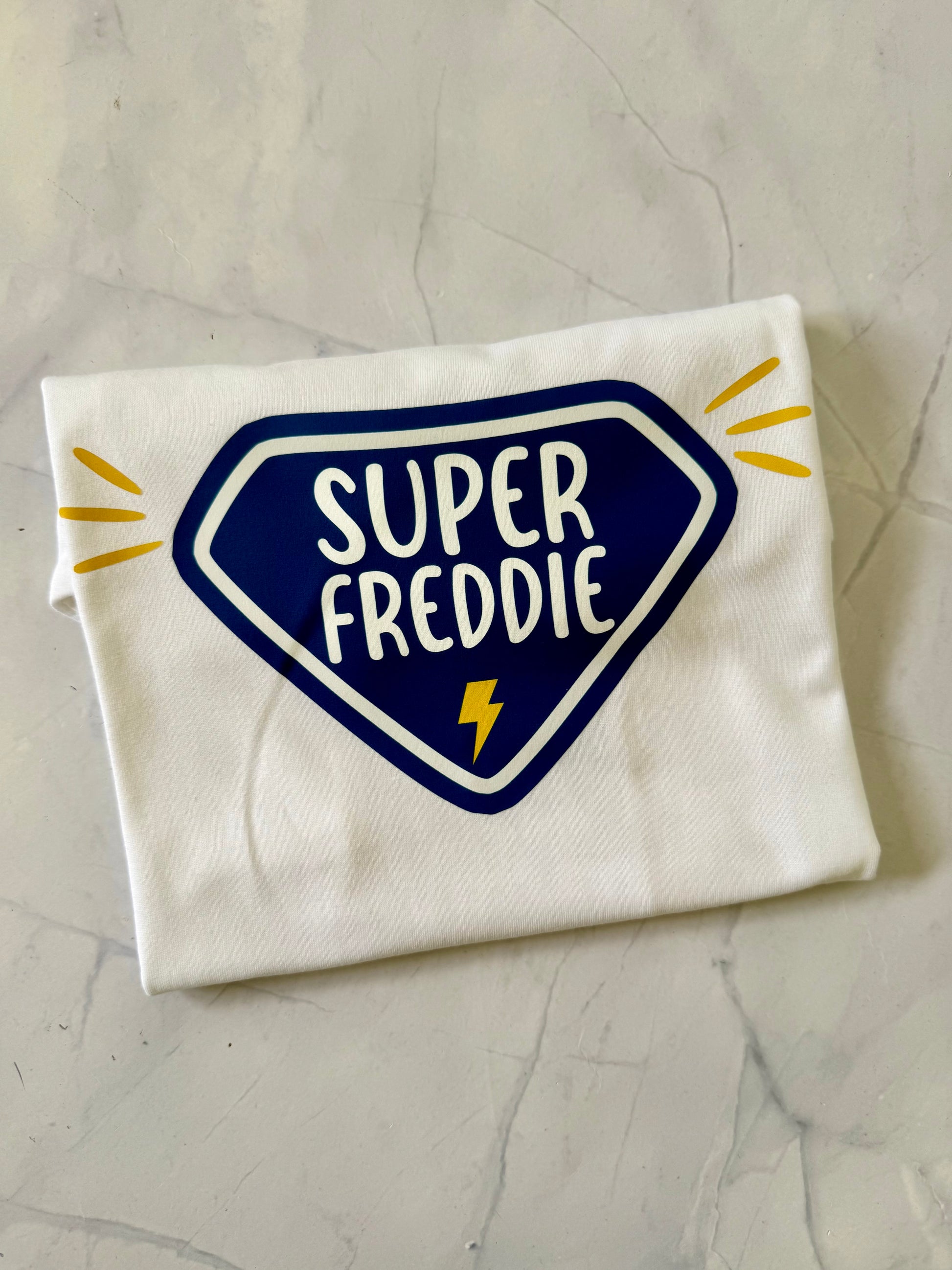 Folded white t-shirt with 'Super Freddie' design on a gray surface
