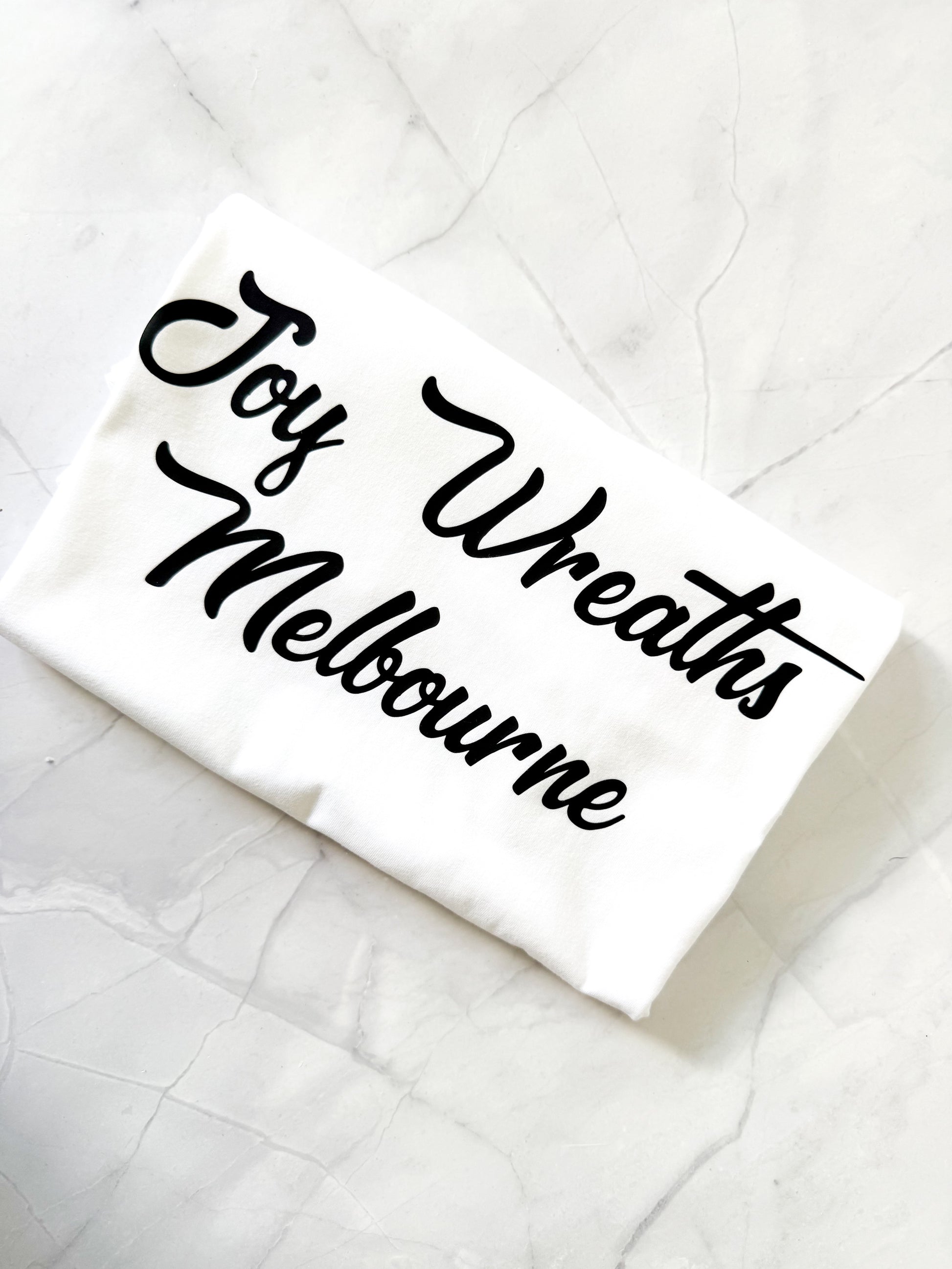White shirt with 'Joy Wreaths Melbourne' text on a textured white surface