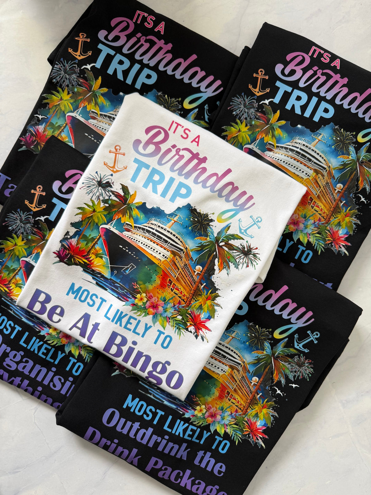 A styled flat-lay of folded graphic T-shirts on a light marble surface. The shirts feature colorful tropical cruise ship illustrations with palm trees, flowers, and anchors, along with humorous birthday-themed text including 'IT'S A Birthday TRIP' and 'MOST LIKELY TO Be At Bingo'.