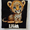 A folded black T-shirt laid flat on a light marble-like surface, featuring a large cartoon leopard cub graphic with blue eyes and the name 'Litia' printed below in white outlined letters.