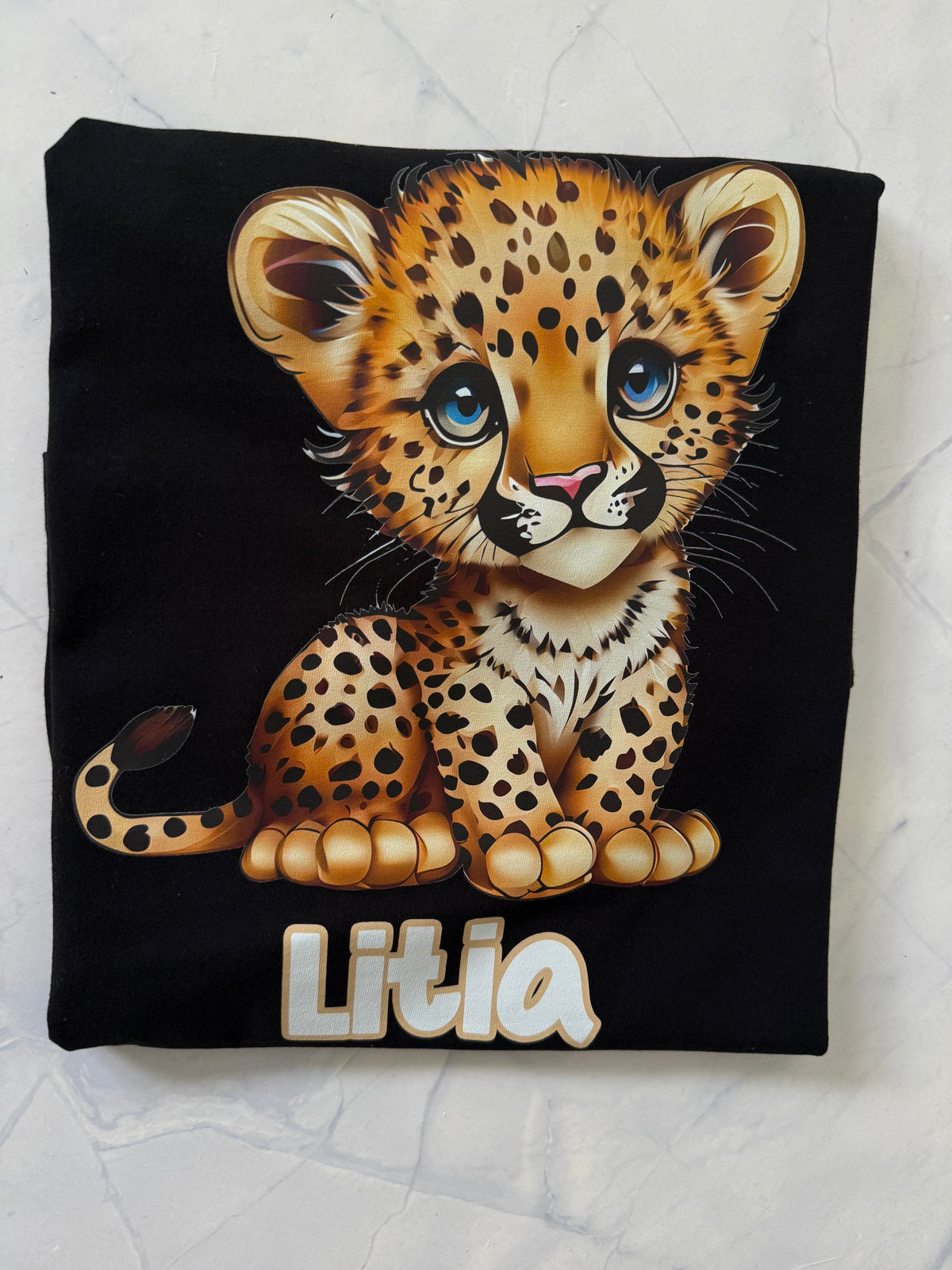 A folded black T-shirt laid flat on a light marble-like surface, featuring a large cartoon leopard cub graphic with blue eyes and the name 'Litia' printed below in white outlined letters.
