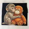 A square black fabric item, likely a bandana or scarf, featuring a detailed illustration of a baby monkey hugging a plush orange toy. The fabric is laid flat on a light gray marble-textured surface.