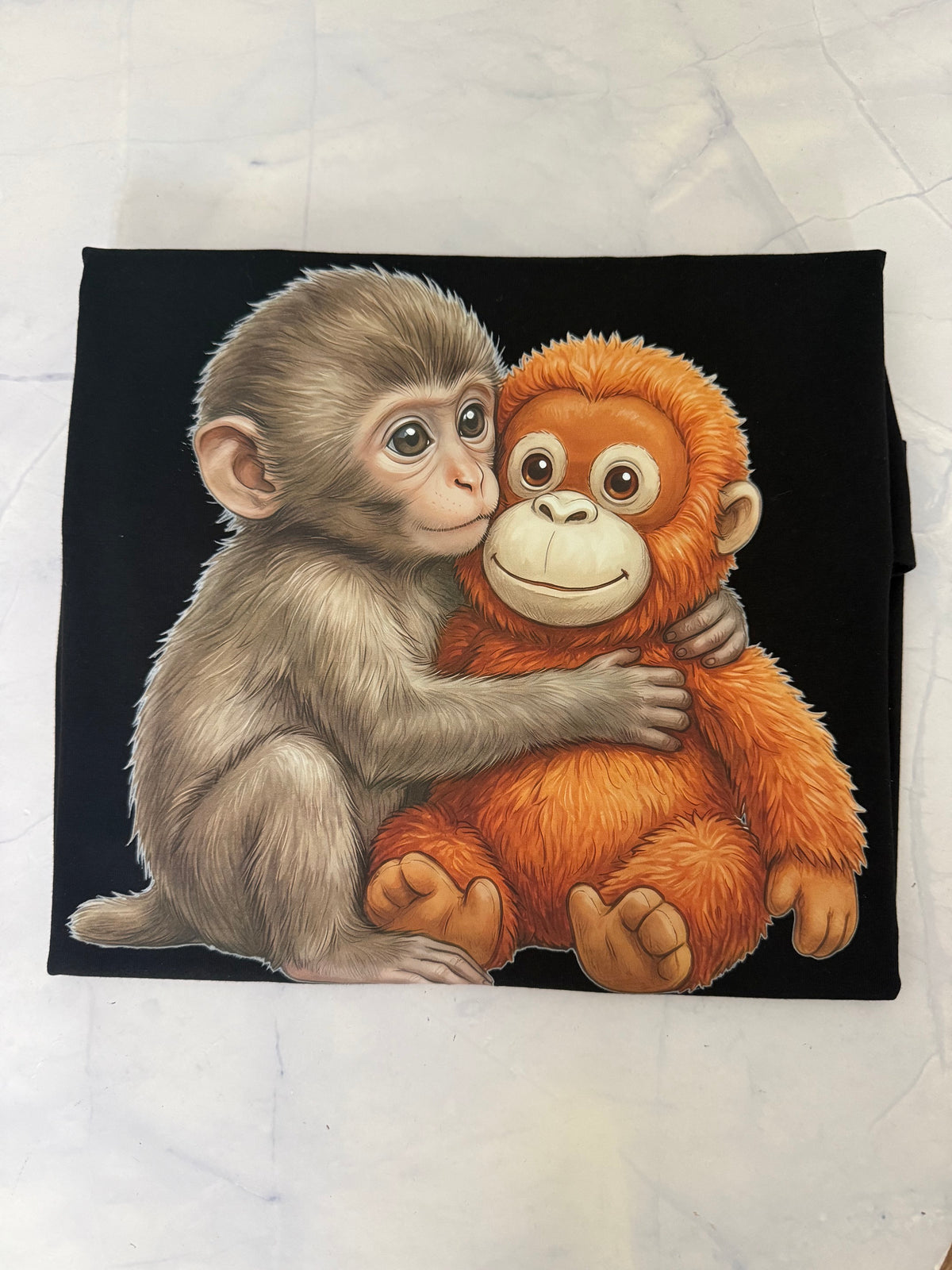 A square black fabric item, likely a bandana or scarf, featuring a detailed illustration of a baby monkey hugging a plush orange toy. The fabric is laid flat on a light gray marble-textured surface.
