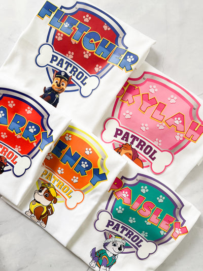 Flat lay of matching Paw Patrol inspired t-shirts for family members with custom names and roles.