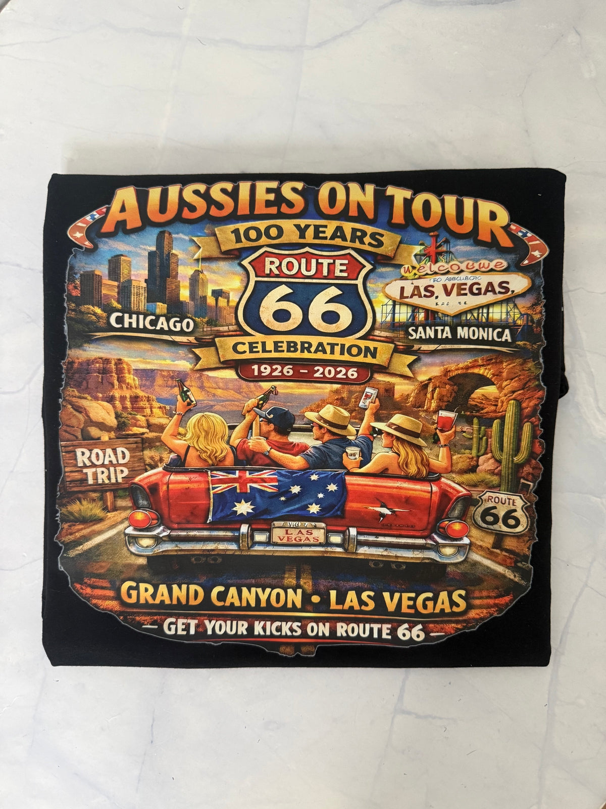 A black graphic t-shirt featuring a vintage-style 'Aussies on Tour 100 Years Route 66 Celebration' design with people in a red car on a desert road trip theme.