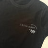 A black short-sleeve crewneck t-shirt with 'TAYLA-MADE PHOTOGRAPHY' and a small leaf graphic printed in white on the left chest. A pink neck label and a white retail tag are visible at the collar.