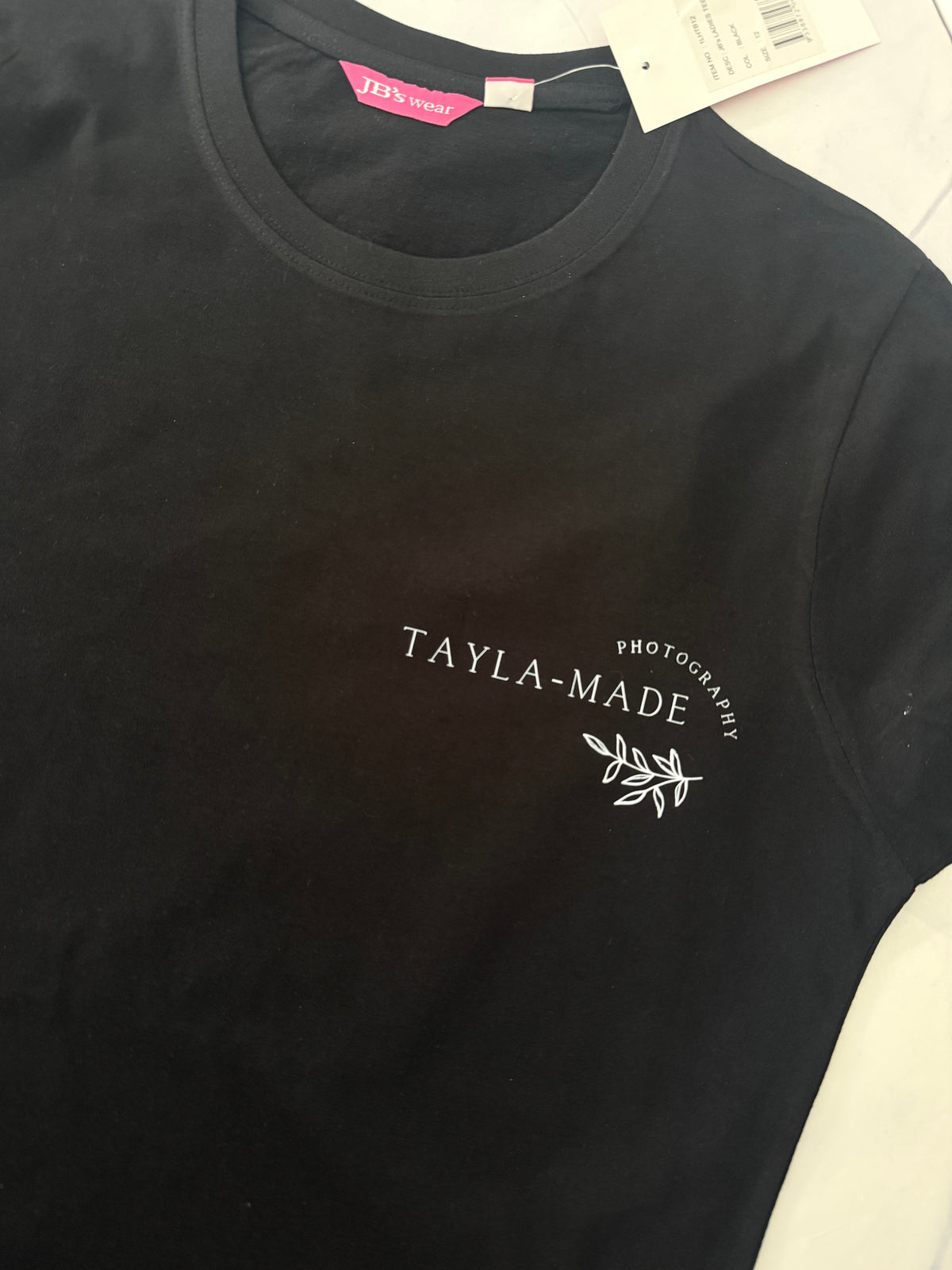 A black short-sleeve crewneck t-shirt with 'TAYLA-MADE PHOTOGRAPHY' and a small leaf graphic printed in white on the left chest. A pink neck label and a white retail tag are visible at the collar.