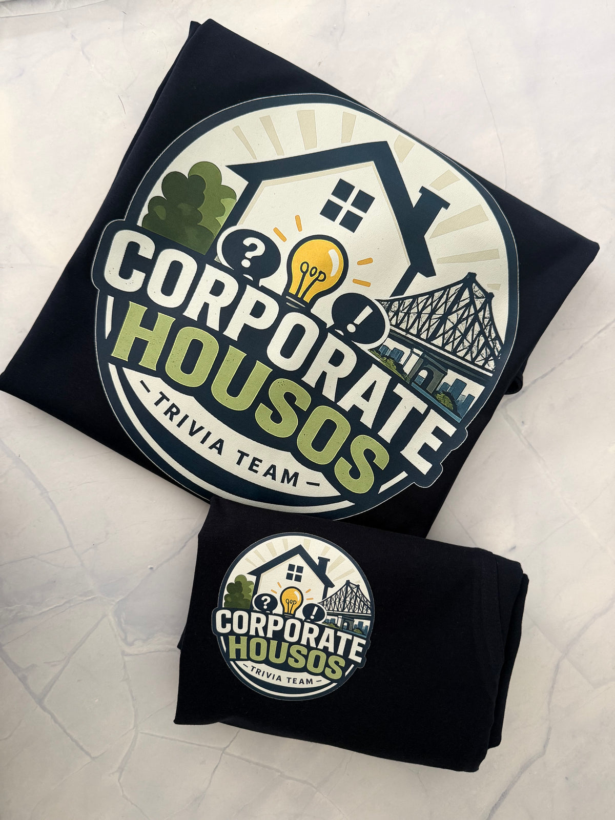 Two folded black T-shirts laid flat on a light textured surface, each printed with a large circular 'Corporate Houses Trivia Team' logo featuring a house, lightbulb, question marks, and bridge graphic.