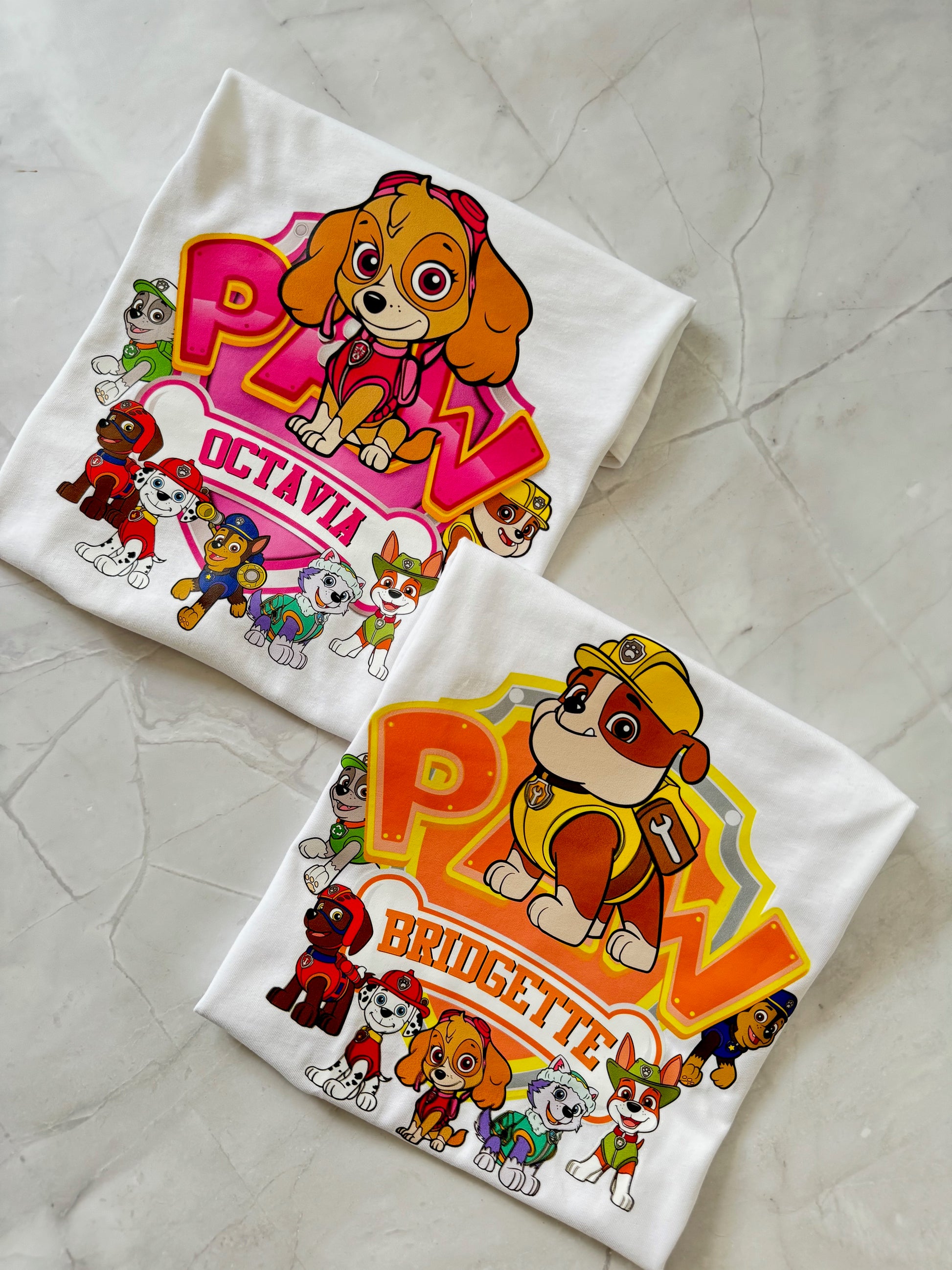 Two personalized PAW Patrol inspired birthday shirts – Skye and Rubble designs – folded on marble background.