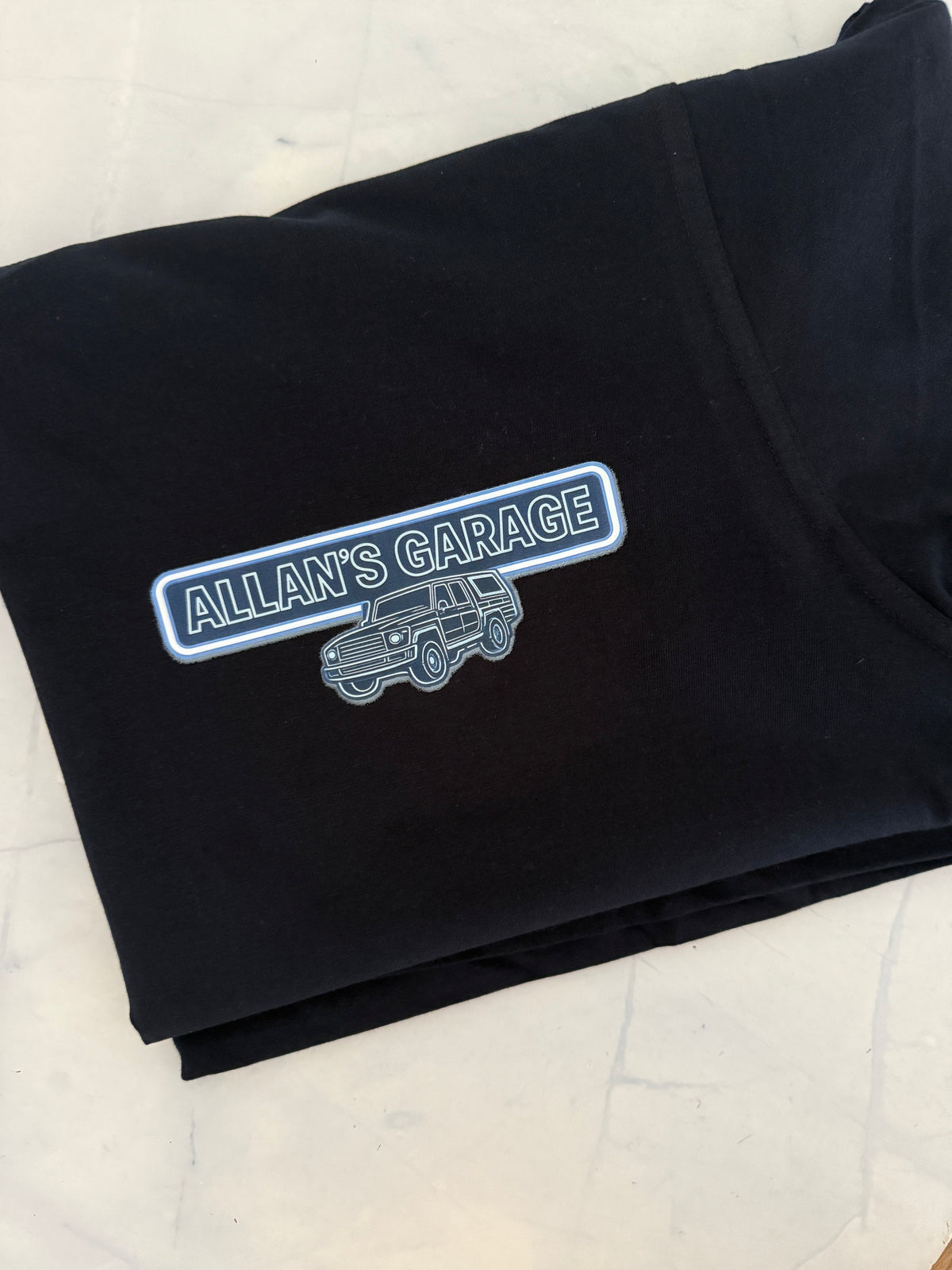 A folded black t-shirt on a light marble surface, featuring a graphic print on the front with a blue and white outlined badge that reads 'ALLAN'S GARAGE' above a silver line-art vehicle illustration.
