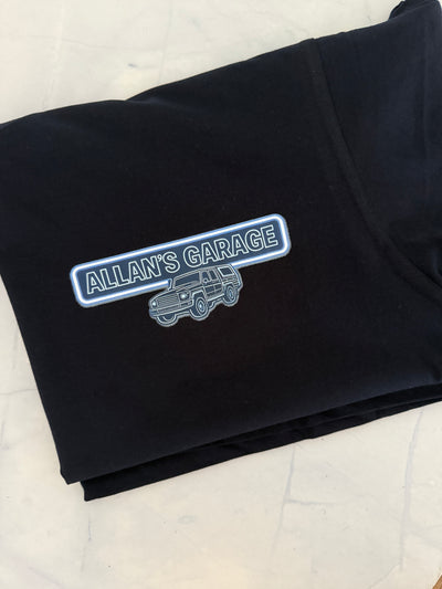 A folded black t-shirt on a light marble surface, featuring a graphic print on the front with a blue and white outlined badge that reads 'ALLAN'S GARAGE' above a silver line-art vehicle illustration.