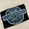 A black folded t-shirt with a circular graphic print reading 'ALLAN'S GARAGE' and 'MY TOOLS / MY RULES' around a car illustration, placed on a light marble surface.