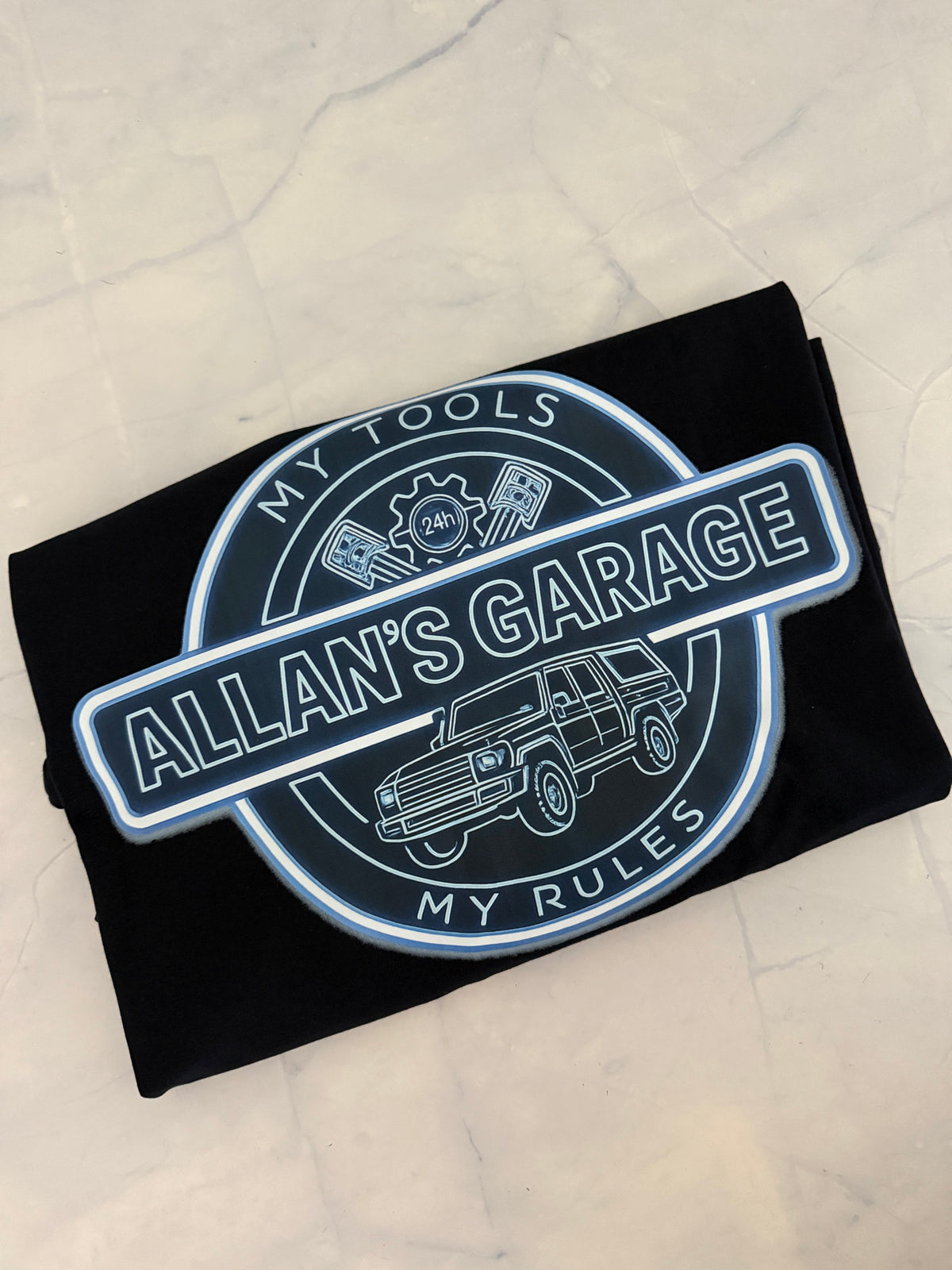 A black folded t-shirt with a circular graphic print reading 'ALLAN'S GARAGE' and 'MY TOOLS / MY RULES' around a car illustration, placed on a light marble surface.