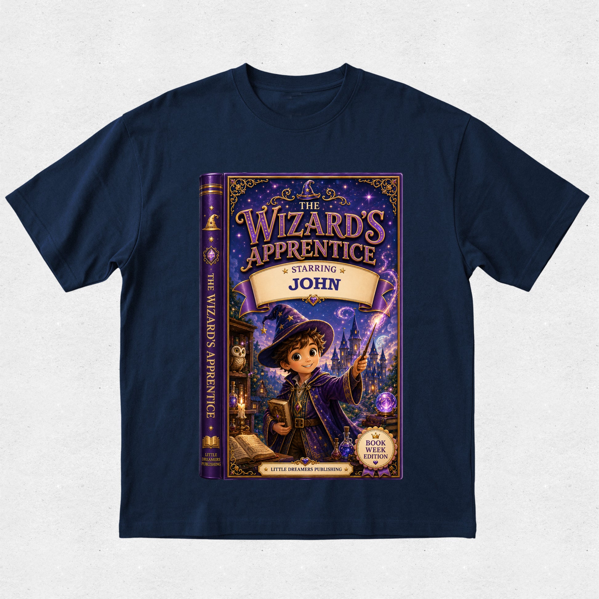 A navy blue short-sleeve t-shirt with a large graphic print of a children's book cover titled 'The Wizard's Apprentice: Starring John' on the front, displayed against a light textured background.