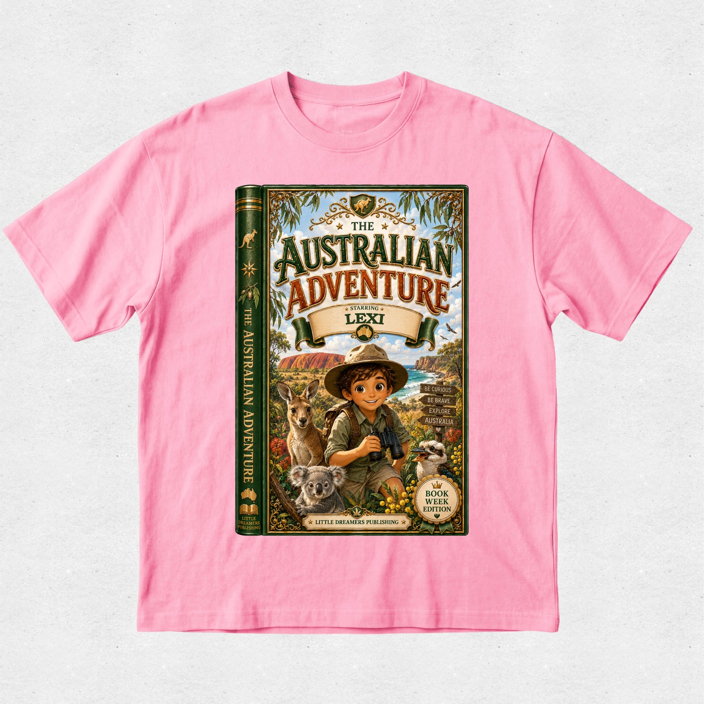 A pink short-sleeve crewneck t-shirt featuring a large graphic print of a children's book titled 'The Australian Adventure' on the front, displayed flat against a light gray textured background.