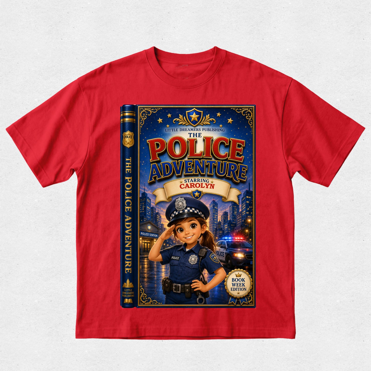 A red short-sleeve crewneck t-shirt featuring a large graphic print of a children's book cover titled 'The Police Adventure: Starting Carolyn'. The graphic depicts a cartoon girl in a police uniform saluting in front of a city skyline with flashing lights, with the book spine visible on the left side of the print.