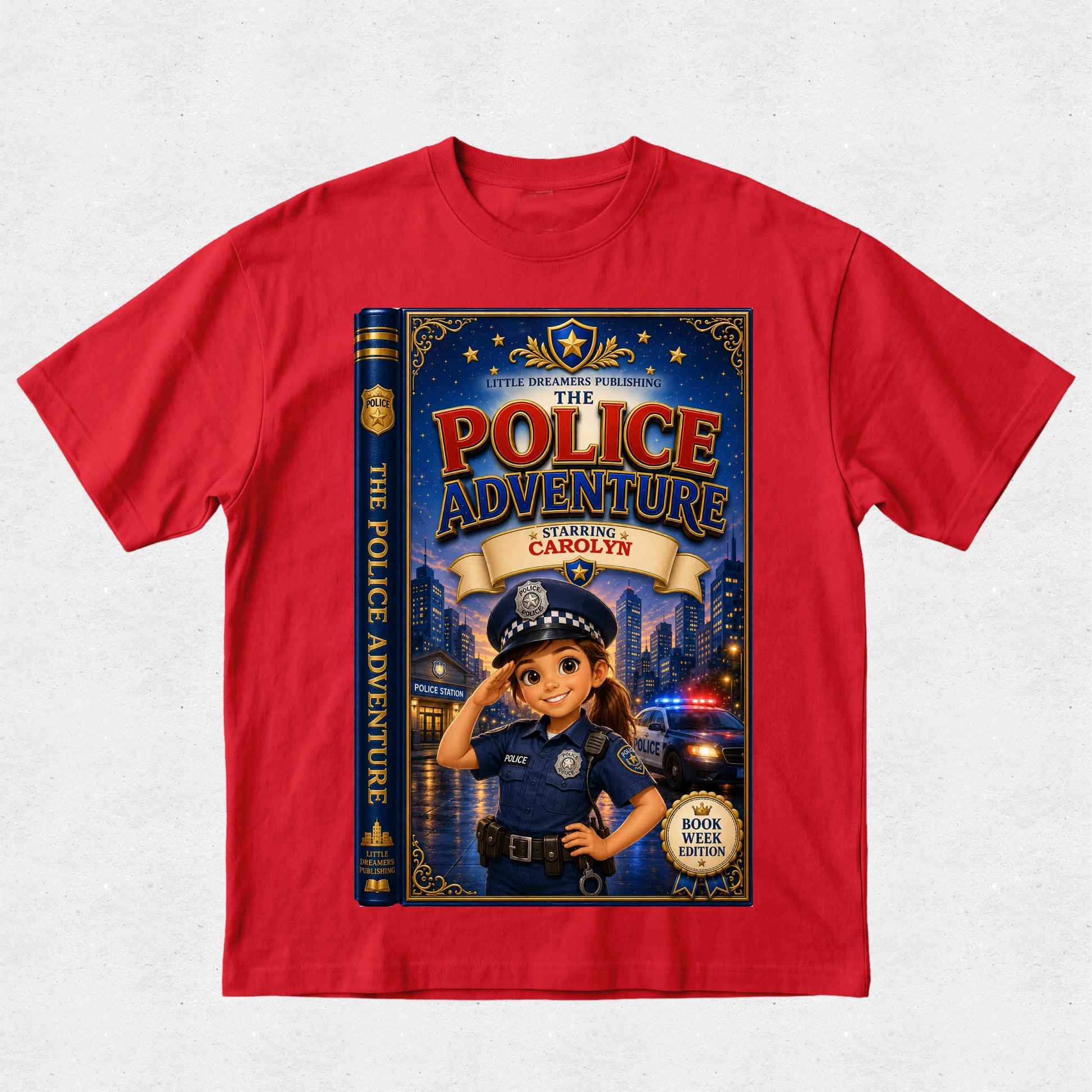 A red short-sleeve crewneck t-shirt featuring a large graphic print of a children's book cover titled 'The Police Adventure: Starting Carolyn'. The graphic depicts a cartoon girl in a police uniform saluting in front of a city skyline with flashing lights, with the book spine visible on the left side of the print.