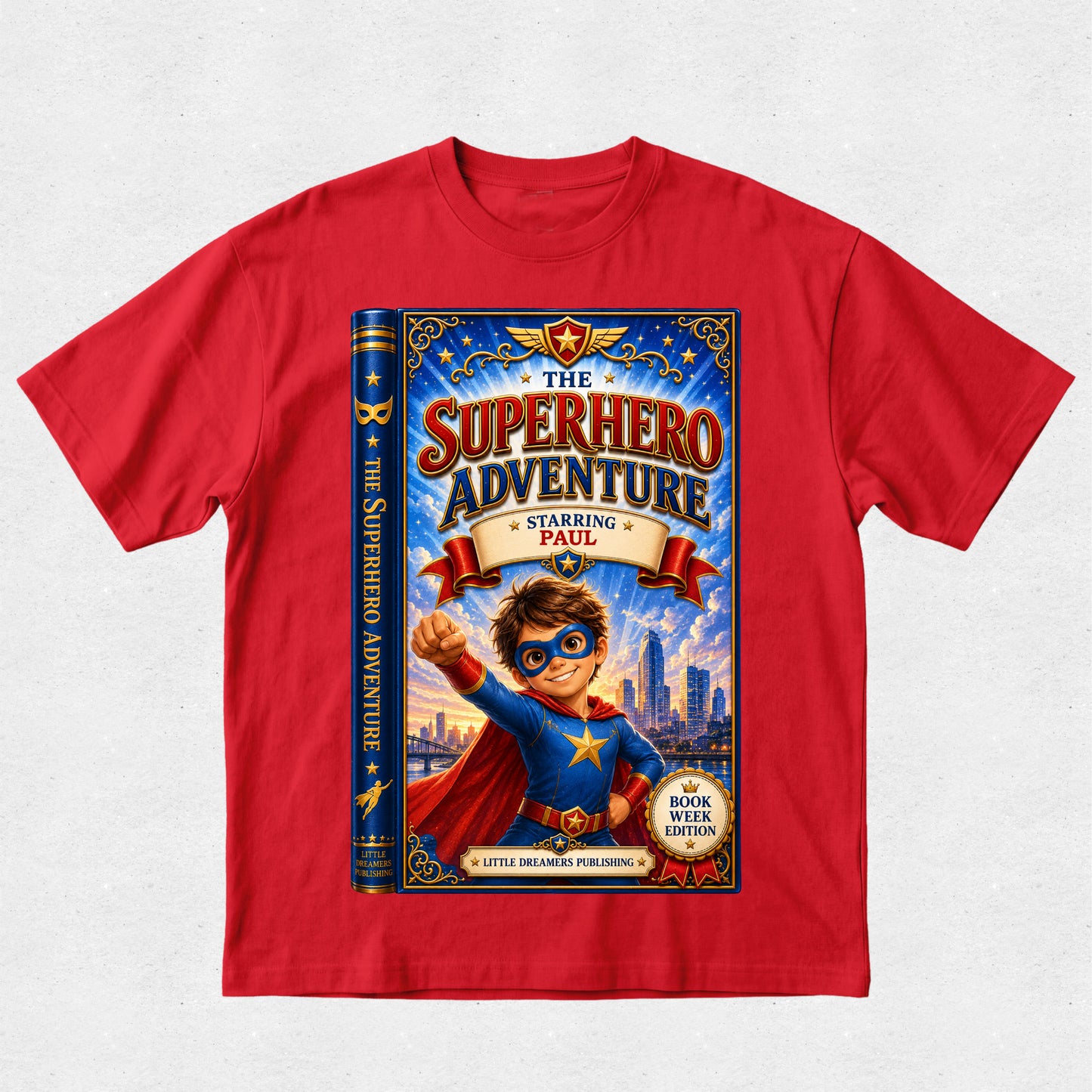 A red short-sleeve crewneck t-shirt featuring a large graphic print of a children's book styled as 'The Superhero Adventure' starring Paul, displayed on a light gray textured background.