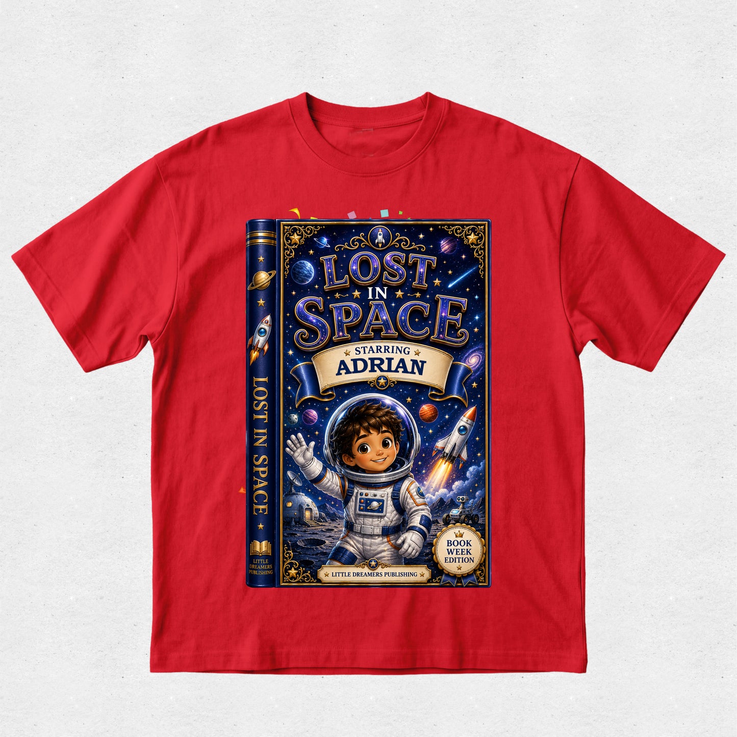 A red short-sleeve crewneck t-shirt featuring a large graphic print of a children's book titled 'Lost in Space' on the front. The graphic shows a dark blue book cover with an astronaut character, stars, and the text 'Starring Adrian'.