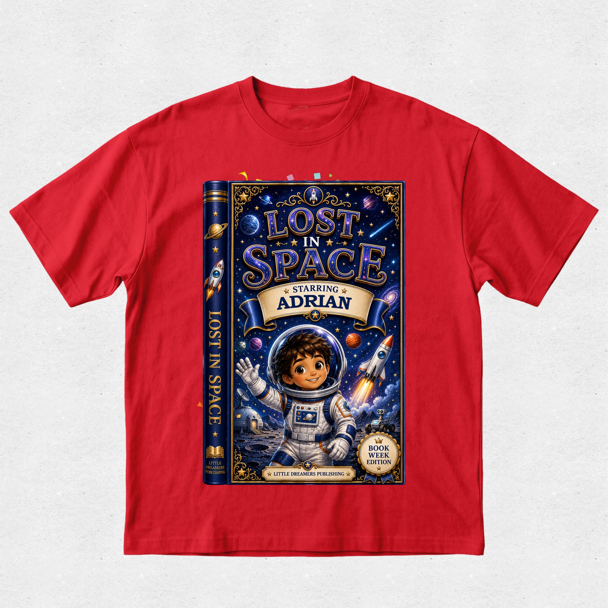 A red short-sleeve crewneck t-shirt featuring a large graphic print of a children's book titled 'Lost in Space' on the front. The graphic shows a dark blue book cover with an astronaut character, stars, and the text 'Starring Adrian'.