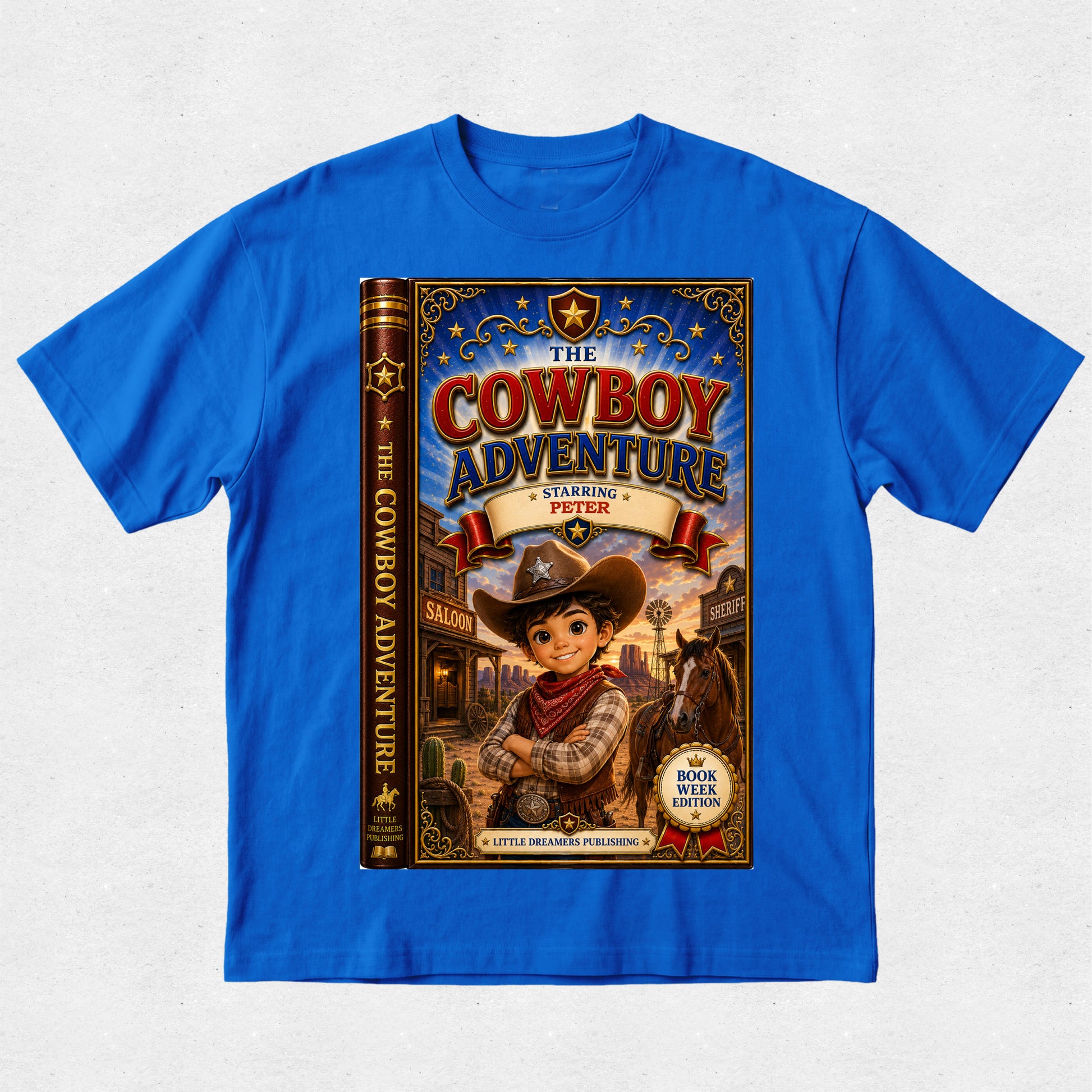 A bright blue short-sleeve crewneck t-shirt featuring a large graphic print of a book cover titled 'The Cowboy Adventure' on the front, displayed flat against a light gray textured background.