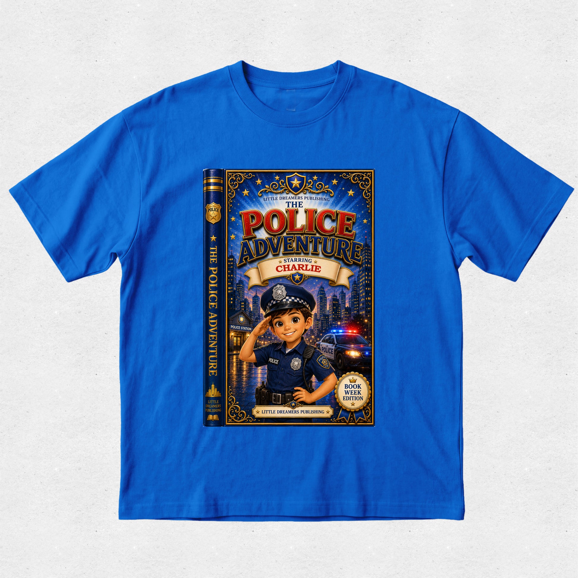 A royal blue short-sleeve t-shirt with a large graphic print of a children's book titled 'The Police Adventure: Charlie' on the front, displayed flat against a light gray textured background.
