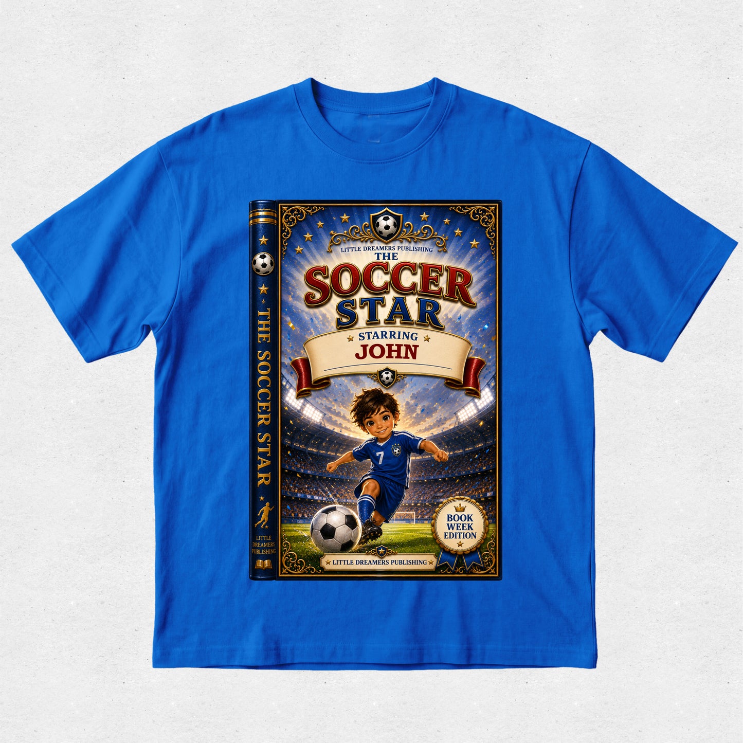 A royal blue short-sleeve t-shirt with a large graphic print of a children's book cover titled 'The Soccer Star Starring John' on the front, displayed flat against a light gray textured background.