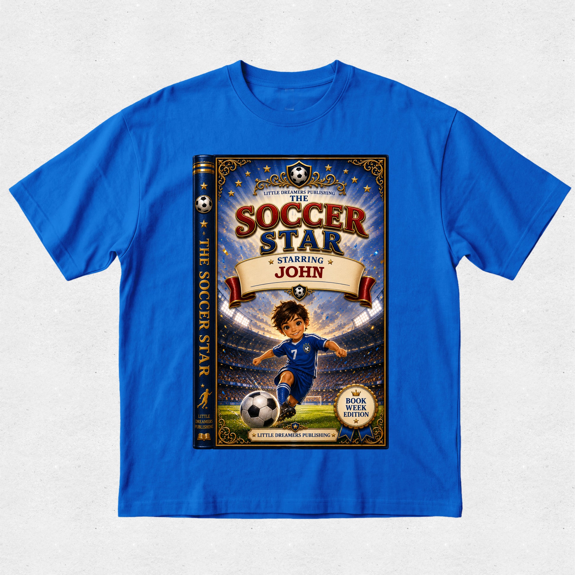 A royal blue short-sleeve t-shirt with a large graphic print of a children's book cover titled 'The Soccer Star Starring John' on the front, displayed flat against a light gray textured background.