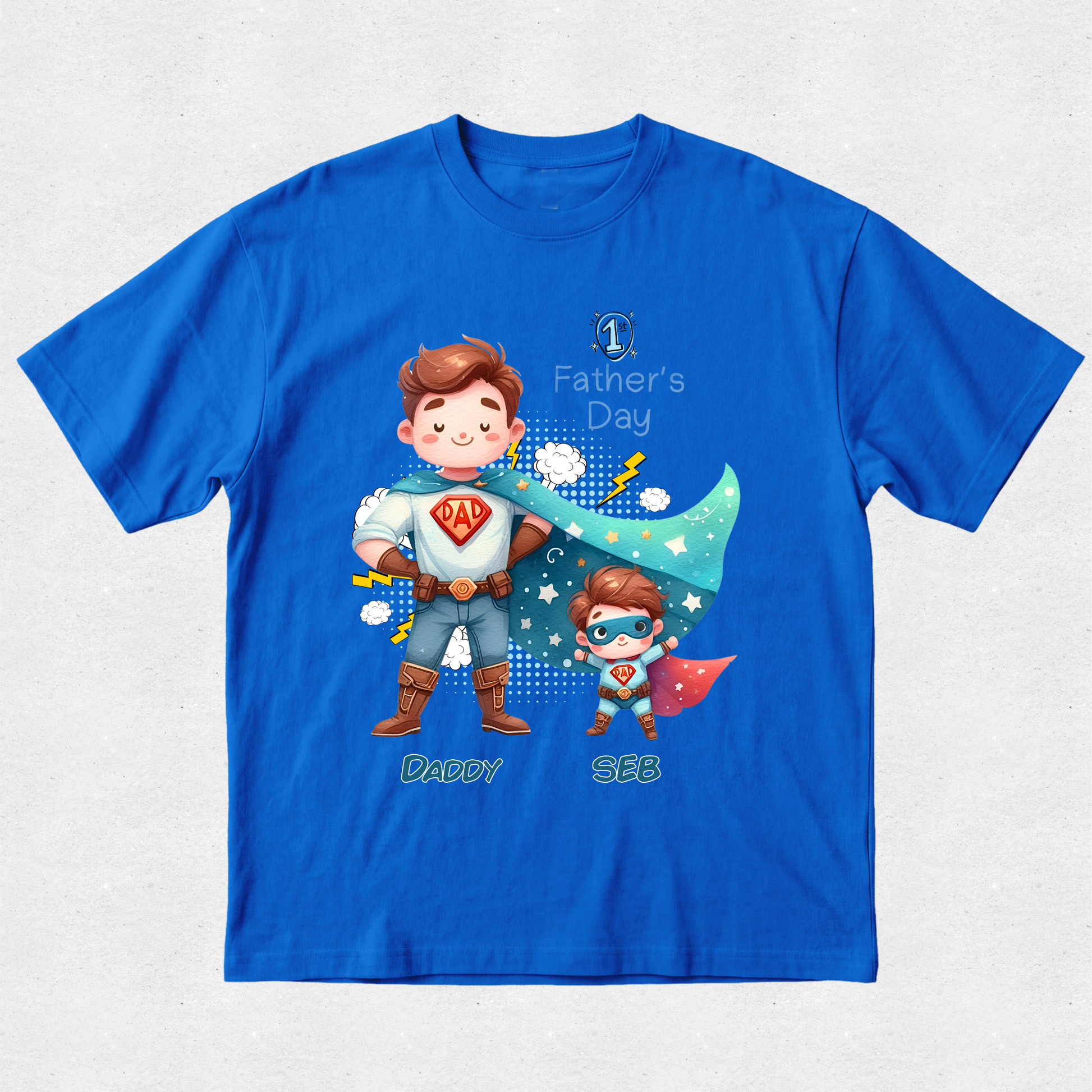 A royal blue short-sleeve t-shirt with a custom cartoon graphic of two superheroes, labeled 'Daddy' and 'Sue', celebrating 'Father's Day'. The shirt is displayed flat against a light gray textured background.