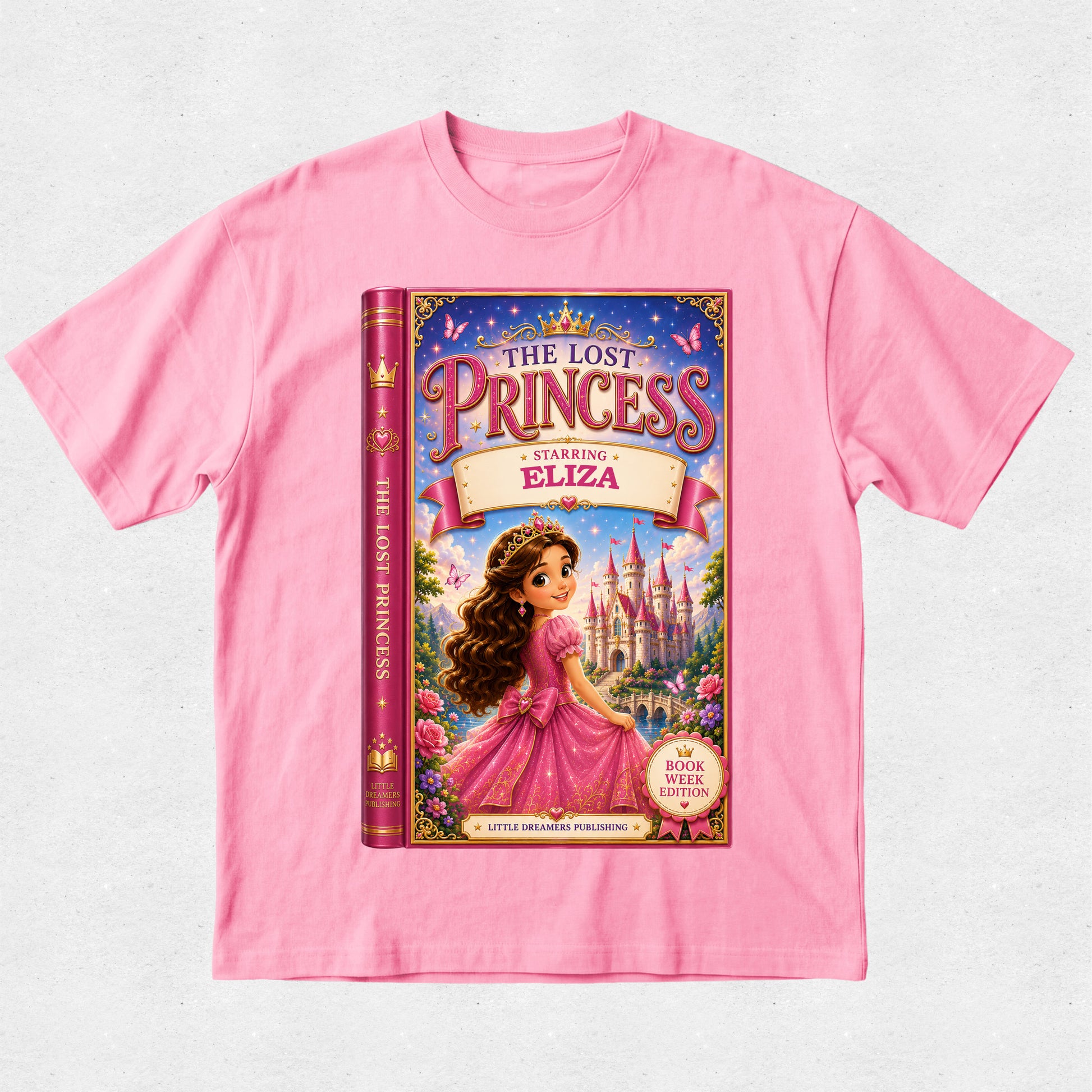 A pink short-sleeve crewneck t-shirt featuring a large graphic print of the book cover for 'The Lost Princess: Starring Eliza' on the front, displayed flat against a light gray textured background.