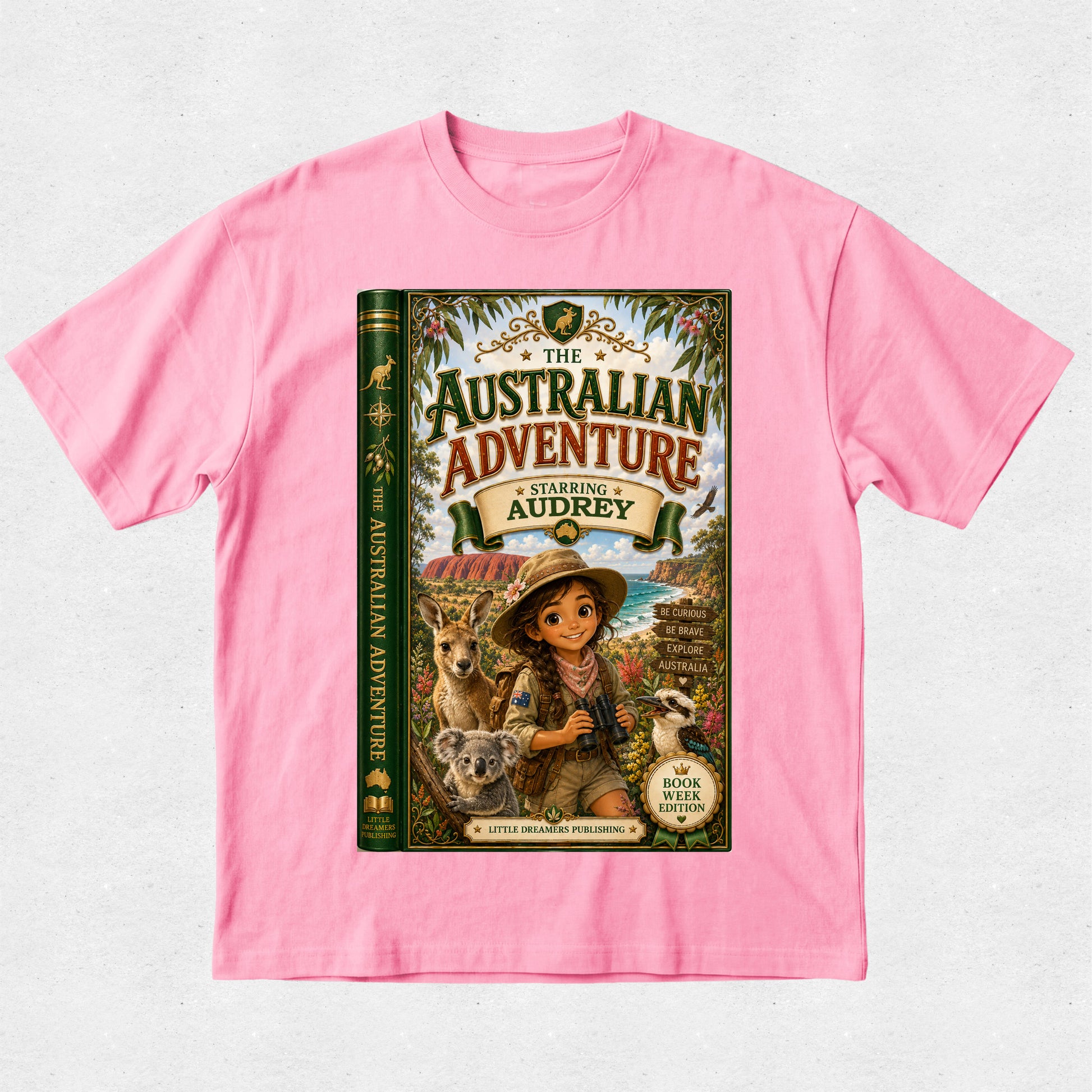 A pink short-sleeve crewneck t-shirt featuring a large graphic print of a book cover titled 'The Australian Adventure Starring Audrey'. The graphic depicts a young girl in a safari hat and vest with a koala and a kangaroo, set against a light gray textured background.