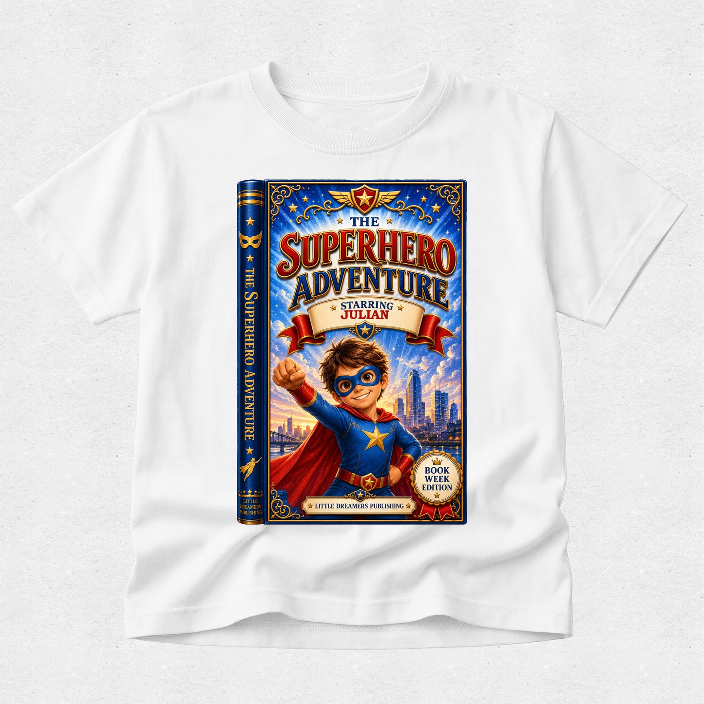 A white kids' t-shirt with a large graphic print of a book cover titled 'The Superhero Adventure: Starring Julian'. The graphic shows a young boy in a superhero costume with a blue cape and mask, standing in front of a city skyline.
