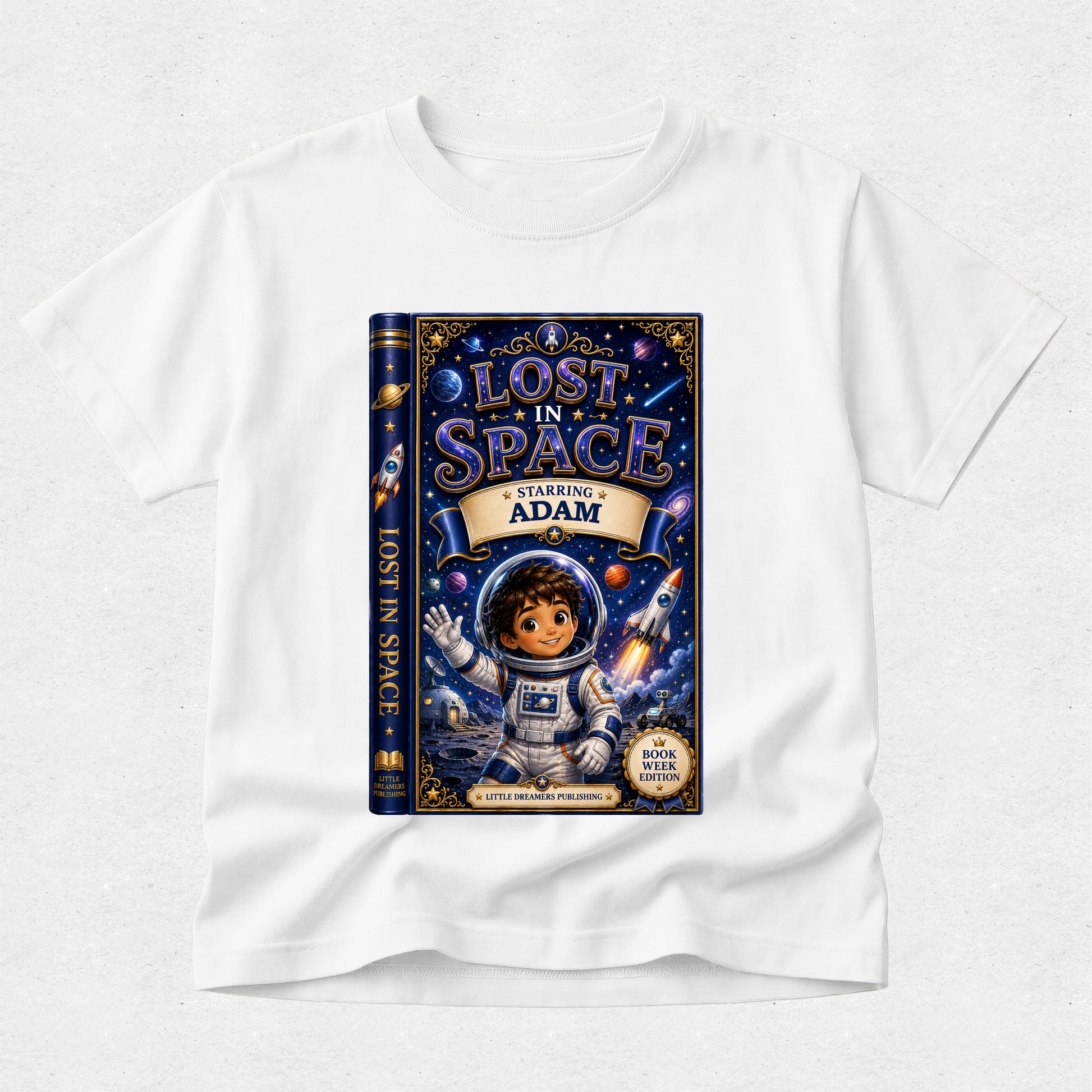 A white short-sleeve crewneck t-shirt featuring a large graphic print of a children's book titled 'Lost in Space: Starring Adam'. The graphic shows a dark blue book cover with an illustration of a young boy in space gear, surrounded by stars, planets, and rockets.