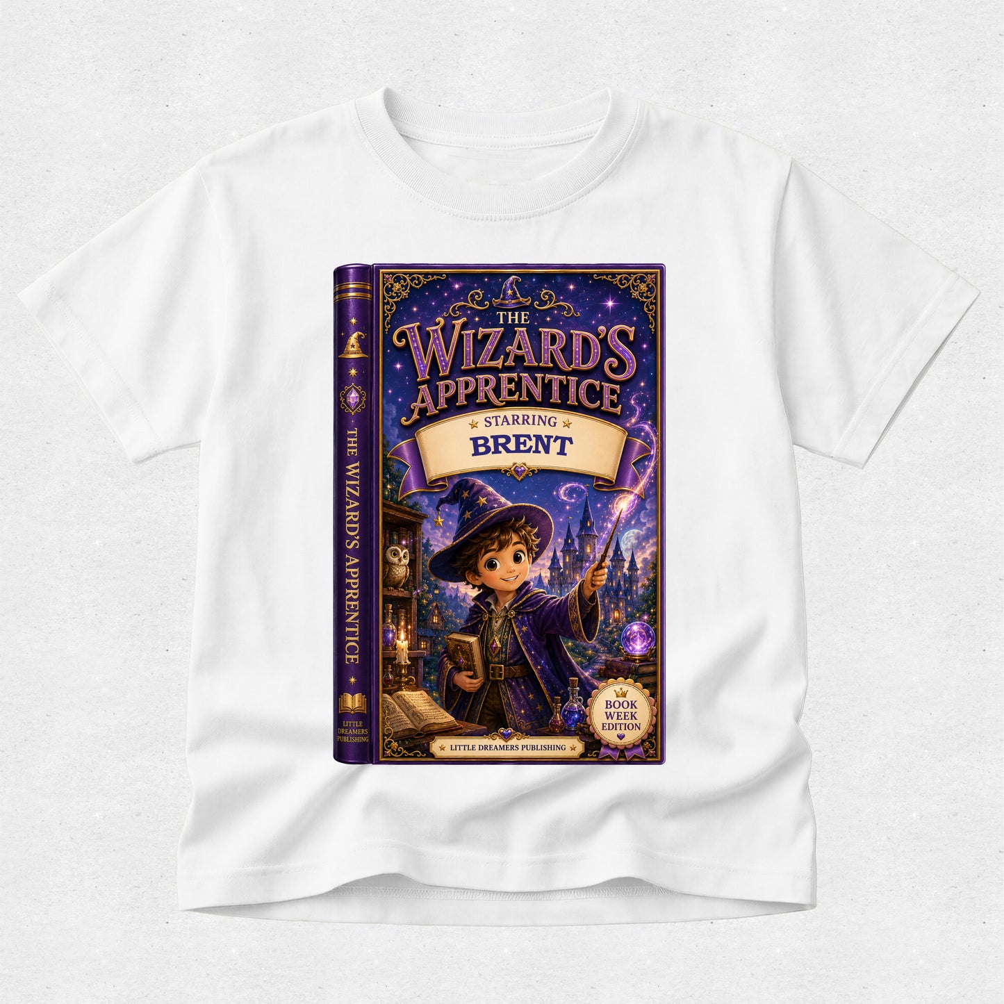 A white short-sleeve crewneck t-shirt featuring a large, colorful graphic print of the book cover for 'The Wizard's Apprentice: Charming Brent' on the front, displayed against a light gray textured background.