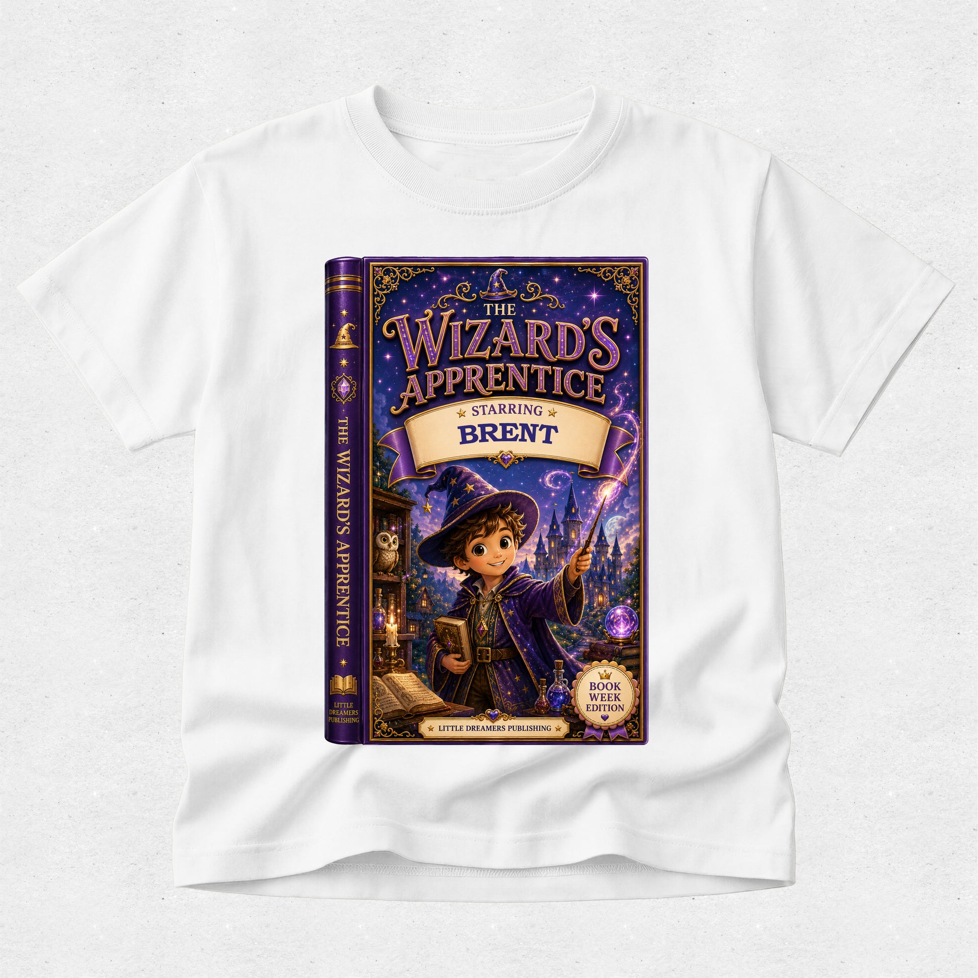 A white short-sleeve crewneck t-shirt featuring a large, colorful graphic print of the book cover for 'The Wizard's Apprentice: Charming Brent' on the front, displayed against a light gray textured background.