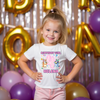 Young girl wearing a 'Birthday Girl' shirt with colorful graphics and text, standing in front of balloons and decorative letters.