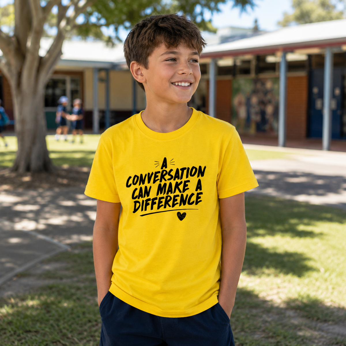 A smiling young boy wearing a bright yellow t-shirt with the black printed text 'A CONVERSATION CAN MAKE A DIFFERENCE' and dark navy shorts, standing outdoors in front of a school building.